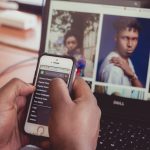 Mãos seguram um smartphone diante de um laptop que exibe imagens, em ambiente interno.