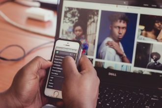 Mãos seguram um smartphone diante de um laptop que exibe imagens, em ambiente interno.