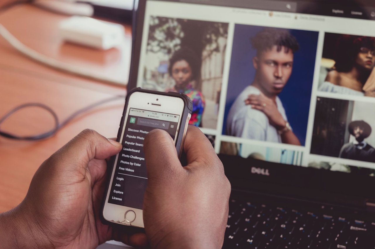 Mãos seguram um smartphone diante de um laptop que exibe imagens, em ambiente interno.