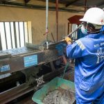 Trabalhador com uniforme azul opera uma mesa vibratória em uma unidade de processamento de minério.