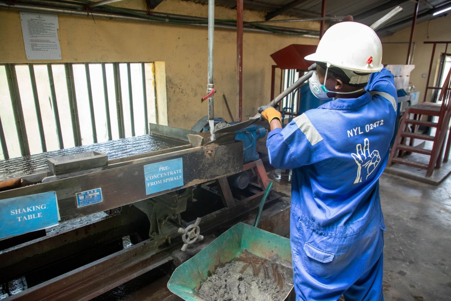 Trabalhador com uniforme azul opera uma mesa vibratória em uma unidade de processamento de minério.