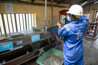 Trabalhador com uniforme azul opera uma mesa vibratória em uma unidade de processamento de minério.