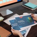 Profissional analisa documentos e gráficos de marketing em uma mesa com computador.